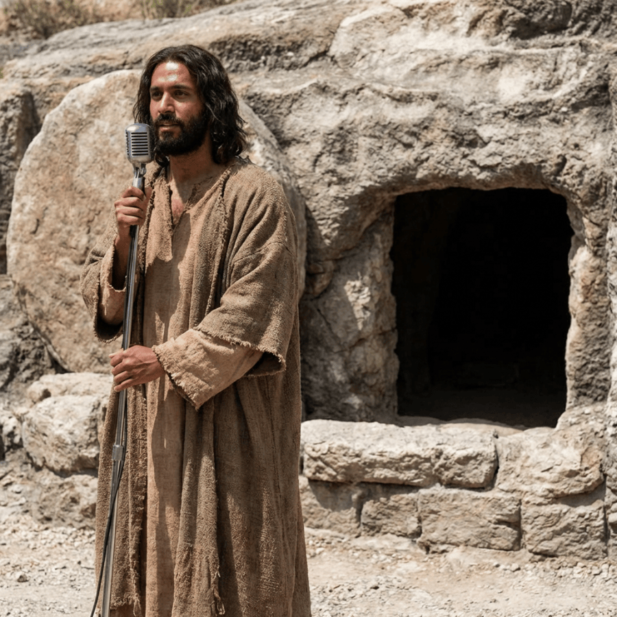 Bearded man in ancient robes holding a microphone in front of a rock tomb.